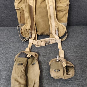Può includere: Uno zaino militare in tela beige con più scomparti, cinghie e tasche. Lo zaino ha schienale e spallacci imbottiti, con tasche aggiuntive e un porta borraccia. Il design suggerisce uno stile vintage o retrò.