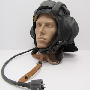 Op de afbeelding: Zwarte leren tankhelm met gehoorbescherming, een kinband en een lange zwarte kabel die eindigt in een meerpins connector. De helm wordt getoond op een mannequinhoofd.