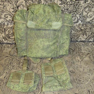 Könnte beinhalten: Ein militärischer Rucksack und zwei Taschen in einem grün-beigen Tarnmuster. Der Rucksack hat einen großen Klappenverschluss und verstellbare Riemen. Die Taschen haben mehrere Fächer.
