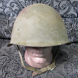 Puede incluir: Un casco militar verde oliva vintage, con una corona redondeada y un borde. El casco tiene una correa para la barbilla y se muestra en una cabeza de maniquí. La superficie del casco muestra signos de desgaste. El fondo es una tela estampada.