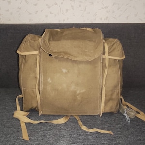 Op de afbeelding: Een beige canvas rugzak met een groot centraal compartiment met een overslagklep, zijzakken en verstelbare stoffen banden. De tas heeft een rechthoekige structuur met zichtbare stiksels en een metalen haakbevestiging aan een van de zijbanden.