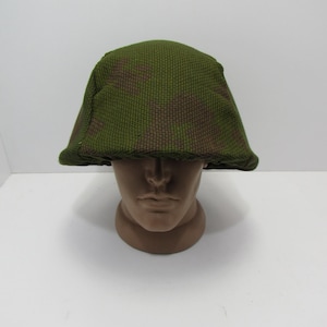 May include: A green and brown camouflage pattern helmet. The helmet is displayed on a mannequin head. The helmet has a rounded shape with a slightly curved brim. The camouflage pattern is a mix of green and brown.