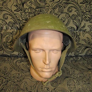 Puede incluir: Un casco militar verde oliva con forma redondeada y barboquejo. El casco se exhibe sobre una cabeza de maniquí contra un fondo estampado.