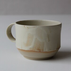 May include: A white ceramic mug with a brown rim and a textured, slightly uneven surface. The mug has a rounded handle.