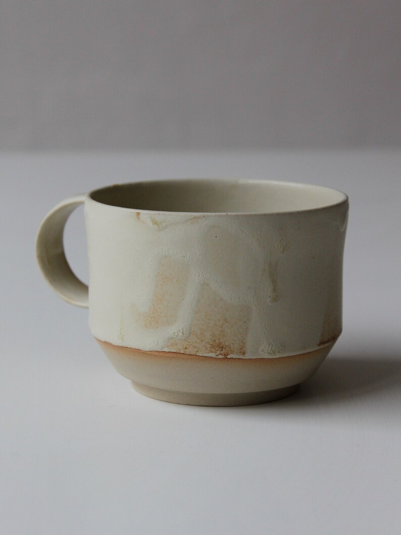 May include: A white ceramic mug with a brown rim and a textured, slightly uneven surface. The mug has a rounded handle.