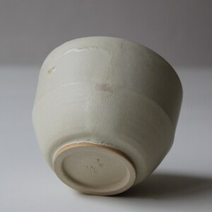 May include: A light beige ceramic bowl with a rounded shape and a slightly textured surface. The bowl has a flat base with a small, circular mark on the bottom.