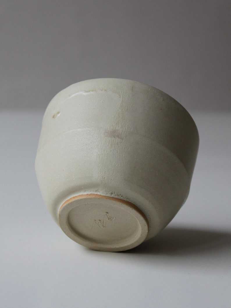 May include: A light beige ceramic bowl with a rounded shape and a slightly textured surface. The bowl has a flat base with a small, circular mark on the bottom.