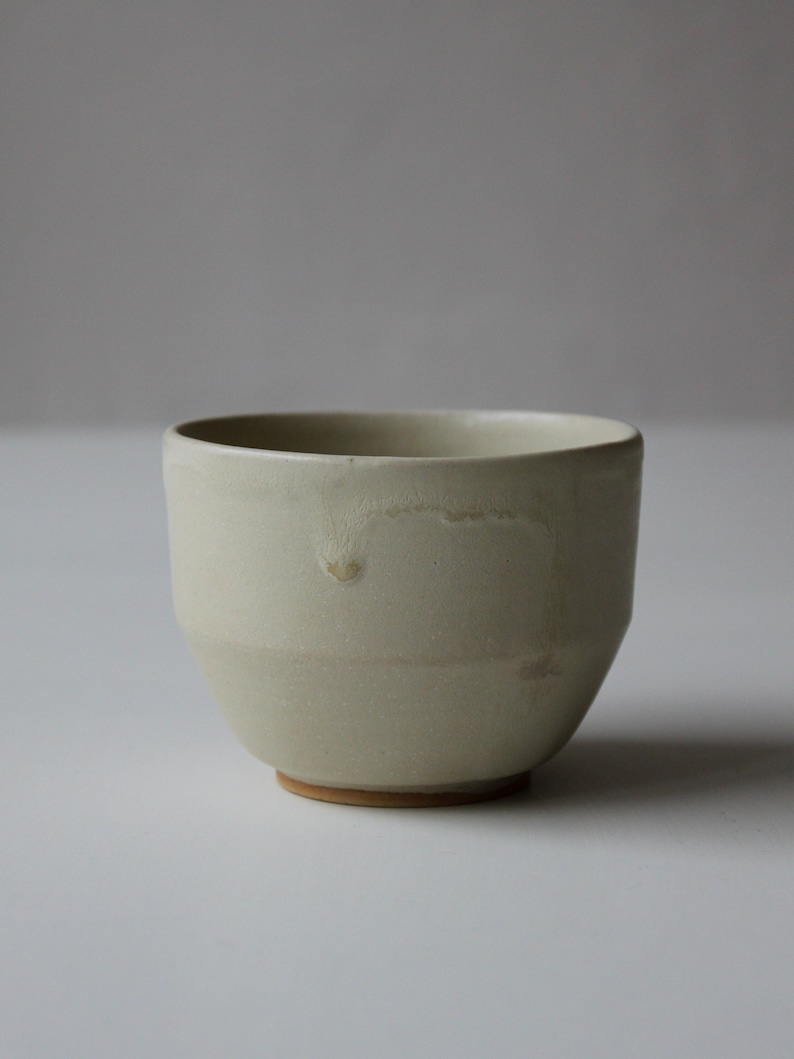 May include: A small, off-white ceramic bowl with a slightly rounded shape and a subtle glaze. The bowl has a slightly textured surface and a light brown rim.