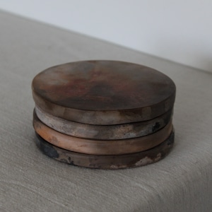 May include: A stack of five round ceramic coasters with a mottled brown, grey, and white glaze. The coasters are stacked on a white linen surface.
