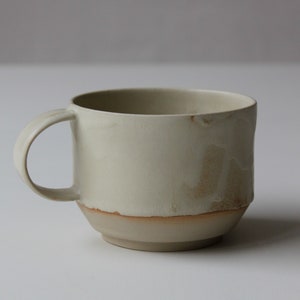 May include: A light beige ceramic mug with a brown rim and a slightly textured surface. The mug has a rounded shape and a handle.