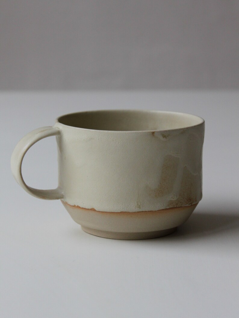 May include: A light beige ceramic mug with a brown rim and a slightly textured surface. The mug has a rounded shape and a handle.