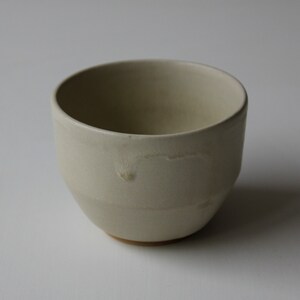 May include: A white ceramic cup with a slightly rounded shape. The cup has a smooth, matte finish and a subtle, uneven texture.