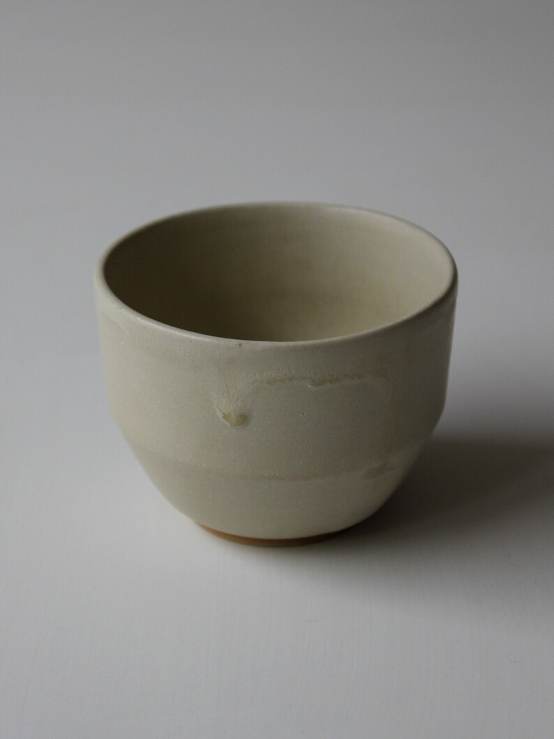 May include: A white ceramic cup with a slightly rounded shape. The cup has a smooth, matte finish and a subtle, uneven texture.