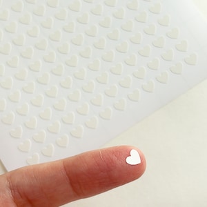 May include: A sheet of white heart-shaped stickers with a single sticker on a finger.