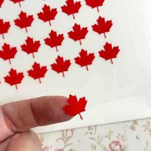 May include: A sheet of red maple leaf stickers. The stickers are arranged in rows on a white background. A finger is holding one of the stickers.