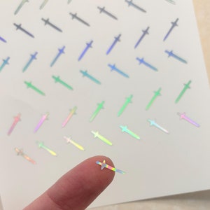 May include: A sheet of iridescent holographic stickers featuring sword shapes in various colors, including blue, green, pink, and yellow. A finger is holding one of the stickers.