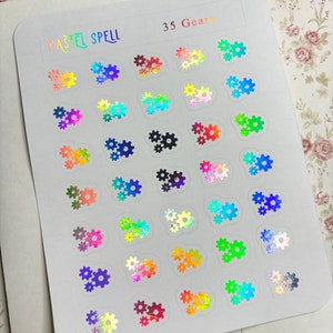 May include: A sheet of holographic stickers featuring gear designs in various colors. The sheet is labeled "PASTEL SPELL" and "35 Gears". The gears have a shiny, iridescent finish, reflecting multiple colors. The stickers are on a white background.