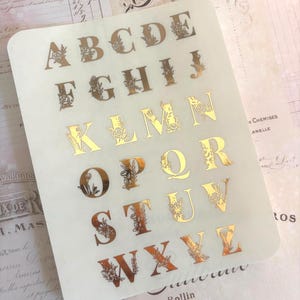 May include: A sheet of gold-colored alphabet stickers, each letter adorned with floral designs. The stickers are on a translucent background, with a vintage-style paper background visible. The letters are in a serif font.