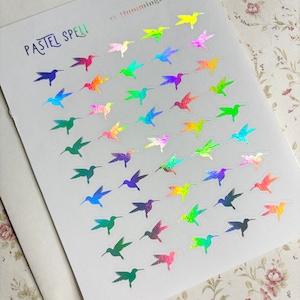 May include: A sheet of stickers featuring 45 iridescent hummingbird designs in various colors. The stickers are arranged on a white background, with the words "PASTEL SPELL" and "45 Hummingbirds" printed on the sheet.