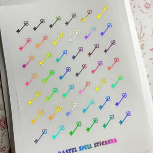 May include: A sheet of pastel holographic key stickers. The stickers are arranged in a diagonal pattern and feature a variety of pastel colors, including pink, blue, green, yellow, and purple. The text "PASTEL SPELL STICKERS" is printed at the bottom of the sheet.