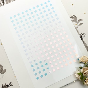 May include: Sheet of star-shaped stickers in a gradient of blue to pink. The stickers are arranged in rows and columns on a white background. The stickers have a holographic, iridescent finish.