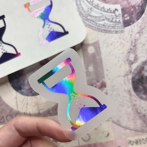 May include: Holographic hourglass sticker with a rainbow sheen. The sticker has a clear border and a design that includes a gradient of colors. The hourglass is filled with a blue liquid. The sticker is held by a hand.