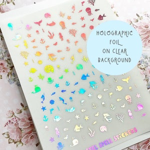 May include: Holographic foil stickers on a clear background, featuring various sea creatures and nautical elements. The stickers include fish, mermaids, anchors, and coral in a rainbow of colors. The text on the image says "Holographic Foil on Clear Background".