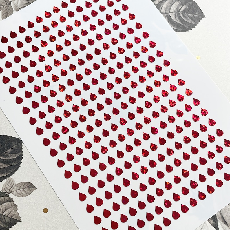 May include: A sheet of red iridescent adhesive stickers in the shape of raindrops. The stickers are arranged in rows on a white background.