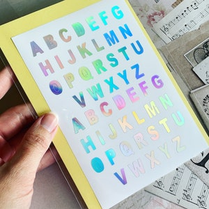 Op de afbeelding: Een vel met regenboogkleurige, iriserende letter stickers. De letters zijn in twee rijen gerangschikt, waarbij de eerste rij begint met A en eindigt met Z, en de tweede rij begint met A en eindigt met Z.