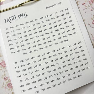 May include: A clipboard with a gold-colored clip holding a white paper with the words "PASTEL SPELL" and a list of numbers from 101 to 200. The background is a floral pattern.