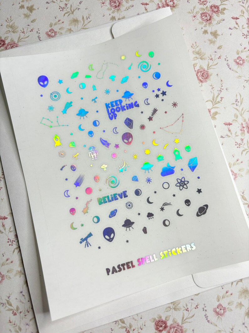 May include: A sheet of iridescent pastel holographic stickers with a variety of space-themed designs, including planets, stars, moons, aliens, and rockets. The sheet also includes the text "Keep Looking Up" and "Believe". The bottom of the sheet reads "Pastel Spell Stickers".