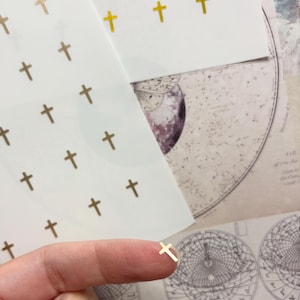 May include: Gold cross stickers on white paper. The stickers are arranged in a grid pattern. A single gold cross sticker rests on a fingertip. The background includes a vintage map and a decorative element.