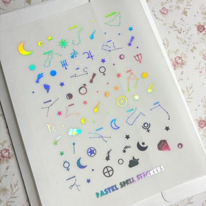 May include: A sheet of iridescent pastel spell stickers featuring various astrological symbols, including constellations, planets, stars, and moons. The text "PASTEL SPELL STICKERS" is printed at the bottom of the sheet.