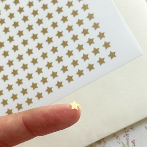 May include: A sheet of gold star stickers with a single star sticker removed and held by a finger. The star stickers are small and have a matte finish.