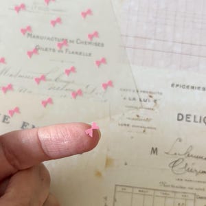 May include: Pink paper bows scattered on a white surface. The bows are small and delicate, and they are perfect for adding a touch of whimsy to any project.