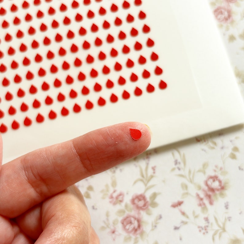 K&ouml;nnte beinhalten: Eine Blatt mit roten, tropfenf&ouml;rmigen Aufklebern. Ein Finger h&auml;lt einen der Aufkleber.