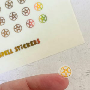 May include: A close-up of a small, round sticker with a gold pentagram design. The sticker is held between fingers, with other stickers and the words "SPELL STICKERS" visible in the background. The stickers have a white background.