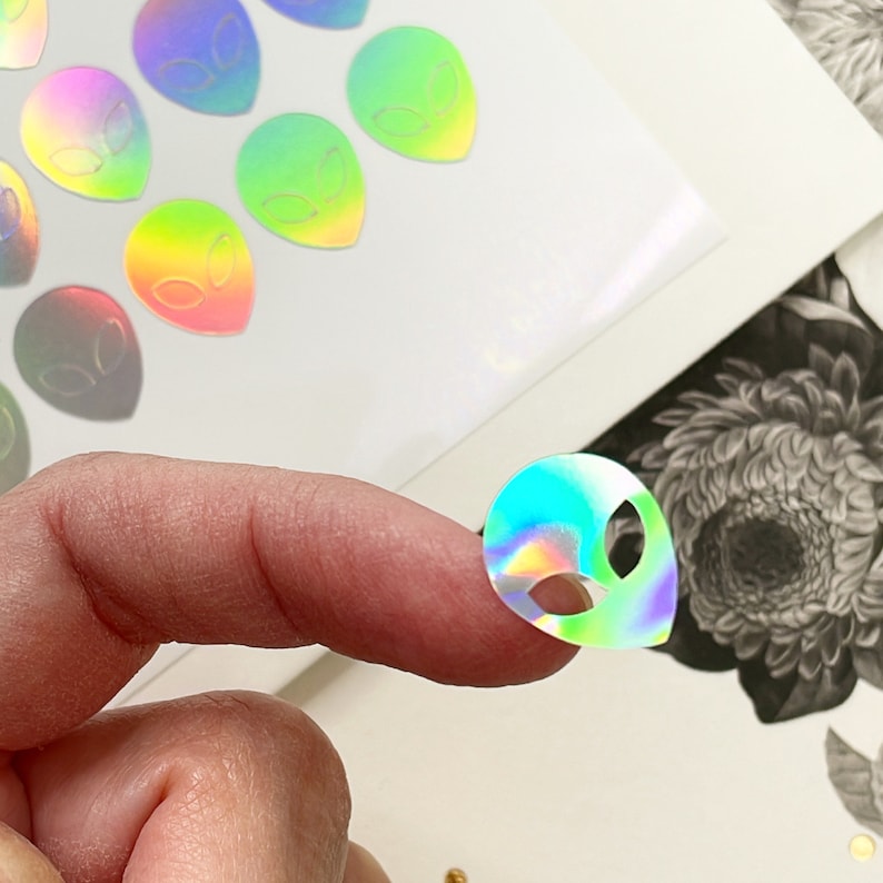 Puede incluir: Una hoja de pegatinas de cabeza de extraterrestre hologr&aacute;ficas iridiscentes. Las pegatinas son de color arco&iacute;ris y tienen un acabado brillante.