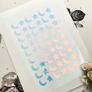 May include: A sheet of iridescent stickers featuring crescent moons and stars in shades of blue, pink, and peach. The stickers are arranged in rows and columns on a clear backing, perfect for scrapbooking or crafts.