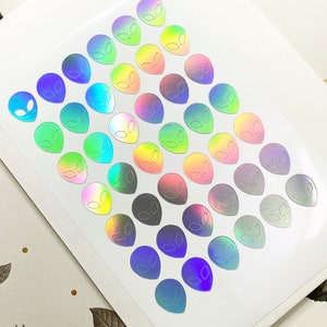 Puede incluir: Una hoja de pegatinas de extraterrestres hologr&aacute;ficas en varios colores arco&iacute;ris. Las pegatinas est&aacute;n dispuestas en filas, siendo la fila superior la m&aacute;s peque&ntilde;a y la fila inferior la m&aacute;s grande. Las pegatinas tienen todas la forma de una cabeza extraterrestre con ojos grandes.