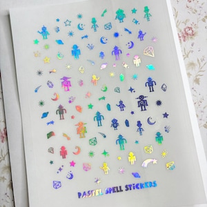 May include: A sheet of iridescent pastel holographic stickers featuring robots, stars, moons, comets, crystals, and other space-themed designs. The sheet is labeled "PASTEL SPELL STICKERS".