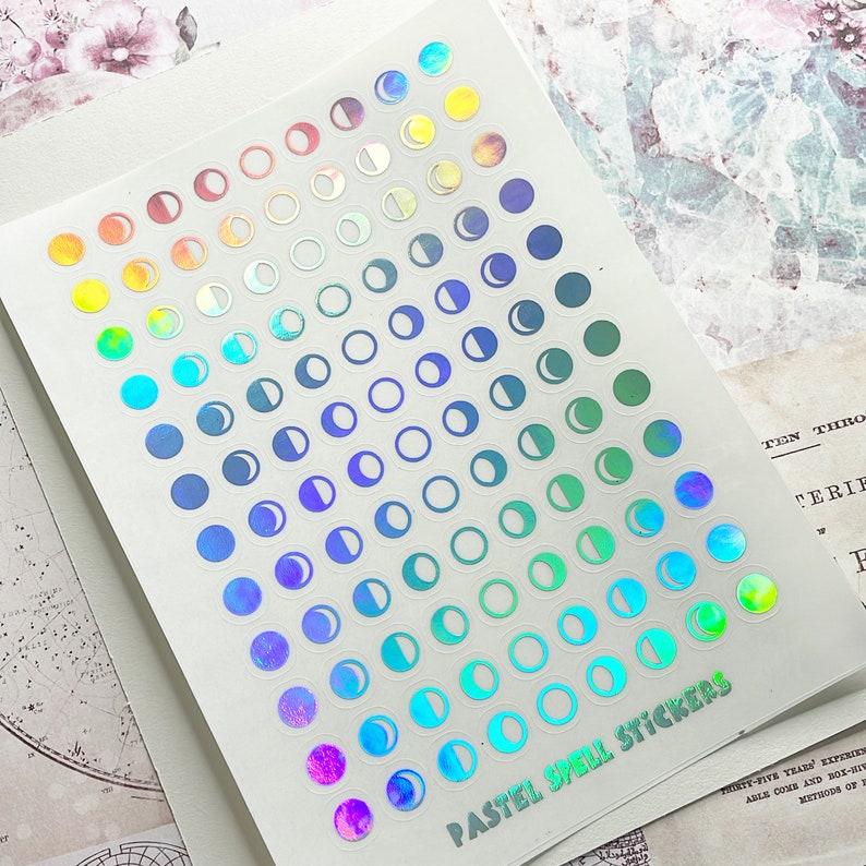 May include: A sheet of iridescent pastel moon phase stickers. The stickers are arranged in a rainbow gradient from red to purple. The sheet has the text "Pastel Spell Stickers" printed on it.