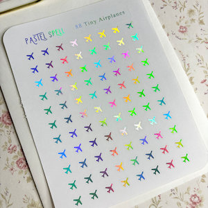 May include: A sheet of 88 iridescent holographic stickers featuring tiny airplane designs in various pastel colors. The sheet is labeled "Pastel Spells 88 Tiny Airplanes".