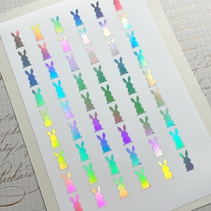 May include: A sheet of holographic stickers featuring a repeating pattern of bunny silhouettes. The stickers display a rainbow of colors, including pink, green, blue, and yellow. The stickers are arranged in neat rows and columns on a white background.