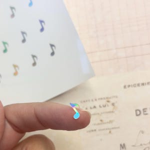 May include: A single iridescent holographic music note sticker. The sticker is on a finger.