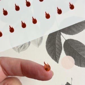 May include: Small, red flame-shaped stickers. The stickers are scattered across a white background with a floral design. One sticker is held on a fingertip, showcasing the size and detail of the design.