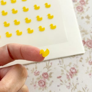 May include: A sheet of yellow rubber duck stickers. The stickers are small and feature a simple outline of a duck. A finger is holding one of the stickers up.