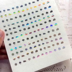May include: A sheet of weather-themed stickers featuring various designs, including clouds, suns, rain, and lightning bolts. Some stickers have a holographic, rainbow-like effect. The stickers are on a white background.