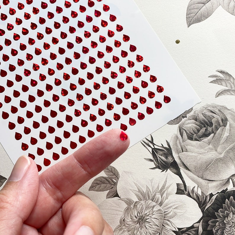 May include: A sheet of red, self-adhesive, glitter-covered, teardrop-shaped stickers. The stickers are arranged in a diagonal pattern on a white sheet of paper. A finger is touching the stickers.