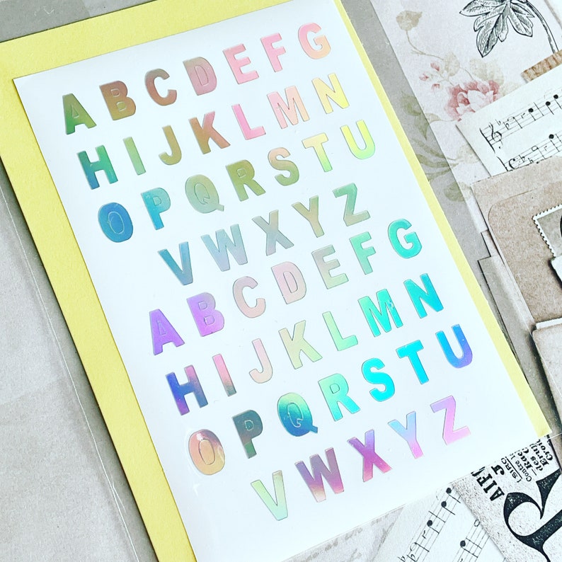 Op de afbeelding: Een vel met regenboogkleurige, iriserende alfabetstickers. De letters zijn in twee rijen gerangschikt, waarbij de eerste rij begint met A en eindigt met G, en de tweede rij begint met H en eindigt met Z.